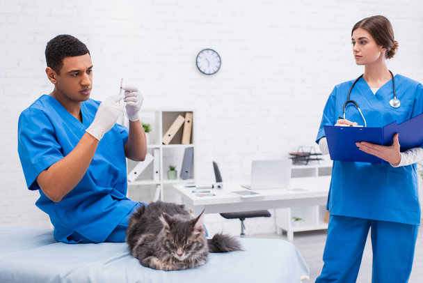 African american veterinarian holding syringe near blurred maine coon and colleague with clipboard in clinic  - Foto, imagen