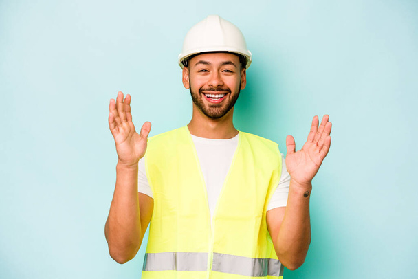 Young laborer hispanic man isolated on blue background receiving a pleasant surprise, excited and raising hands. - 写真・画像