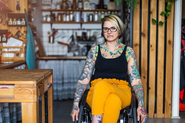 Πορτρέτο μιας νεαρής γυναίκας με τατουάζ σε αναπηρικό καροτσάκι στο σπίτι - Φωτογραφία, εικόνα