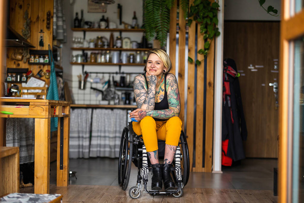 Portret van een jonge getatoeëerde vrouw in een rolstoel thuis - Foto, afbeelding