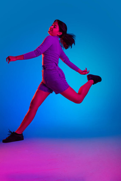 Full-length portrait of young girl cheerfully jumping isolated over gradient blue pink background in neon light - Foto, Imagem