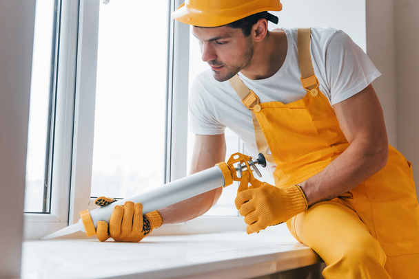 Handyman in yellow uniform works with glue for window indoors. House renovation conception. - Φωτογραφία, εικόνα