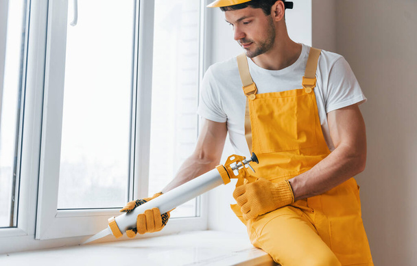 Handyman in yellow uniform works with glue for window indoors. House renovation conception. - Φωτογραφία, εικόνα