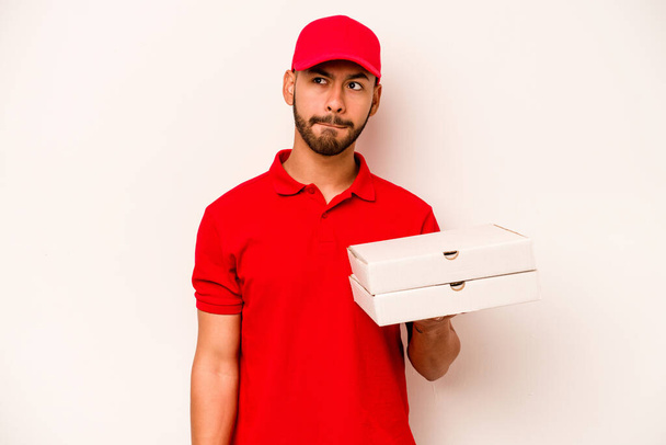 Young hispanic delivery man holding pizzas isolated on white background confused, feels doubtful and unsure. - Photo, image
