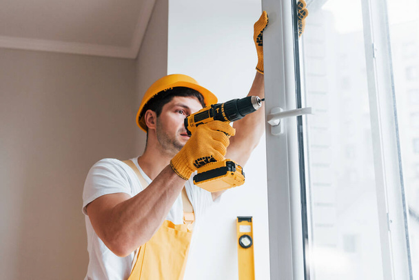 Handyman in yellow uniform installs new window by using automatic screwdriver. House renovation conception. - Фото, изображение