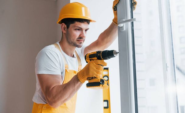 Handyman in yellow uniform installs new window by using automatic screwdriver. House renovation conception. - Zdjęcie, obraz