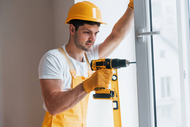 Handyman in yellow uniform installs new window by using automatic screwdriver. House renovation conception. - 写真・画像