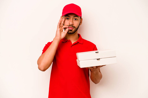 Fiatal spanyol futár, fehér alapon izolált pizzákat tart, ujjaival az ajkain titkot tart.. - Fotó, kép