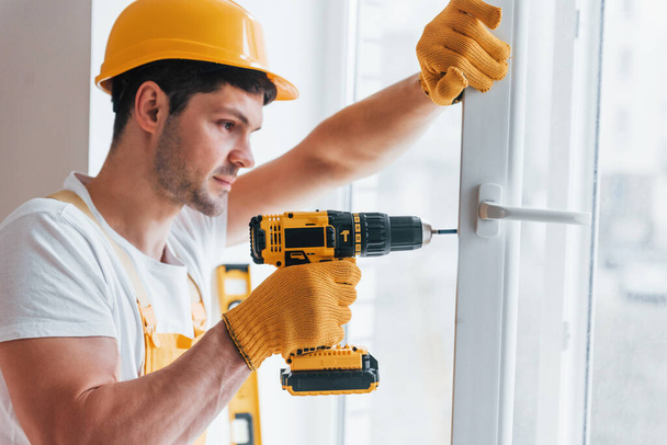 Handyman in yellow uniform installs new window by using automatic screwdriver. House renovation conception. - 写真・画像