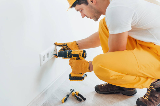 Handyman in yellow uniform works with electricity and installing new socket by using automatic screwdriver. House renovation conception. - Foto, Bild