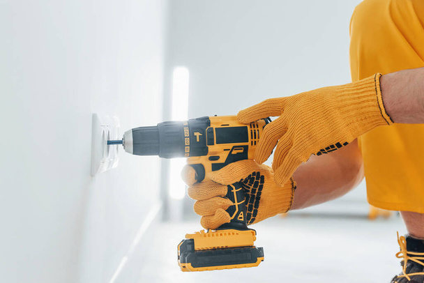 Handyman in yellow uniform works with electricity and installing new socket by using automatic screwdriver. House renovation conception. - Photo, Image