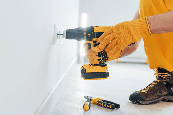 Handyman in yellow uniform works with electricity and installing new socket by using automatic screwdriver. House renovation conception. - Valokuva, kuva