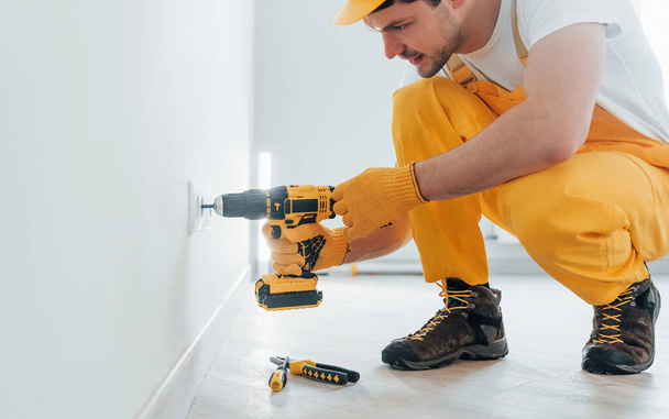 Handyman in yellow uniform works with electricity and installing new socket by using automatic screwdriver. House renovation conception. - Foto, Imagem