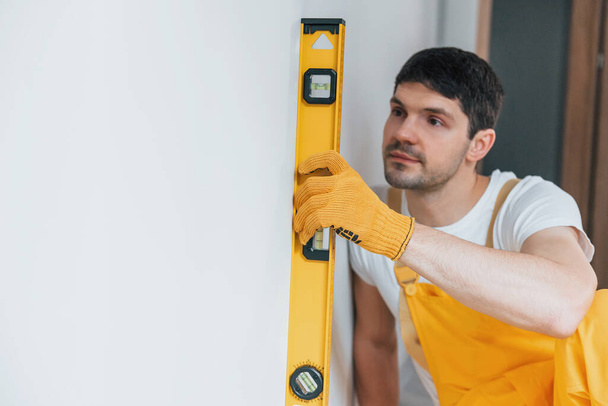 Handyman in yellow uniform checks levels of wall. House renovation conception. - Photo, Image