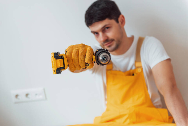 Handyman in yellow uniform standing against white wall with automatic screwdriver. House renovation conception. - Фото, изображение
