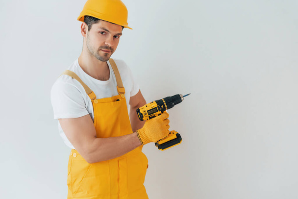 Handyman in yellow uniform standing against white wall with automatic screwdriver. House renovation conception. - Fotó, kép