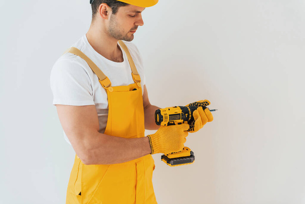 Handyman in yellow uniform standing against white wall with automatic screwdriver. House renovation conception. - Valokuva, kuva