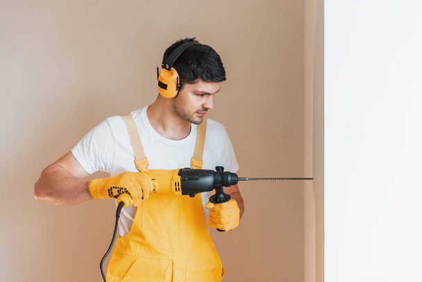 Handyman in yellow uniform works indoors by using hammer drill. House renovation conception. - Foto, Imagen