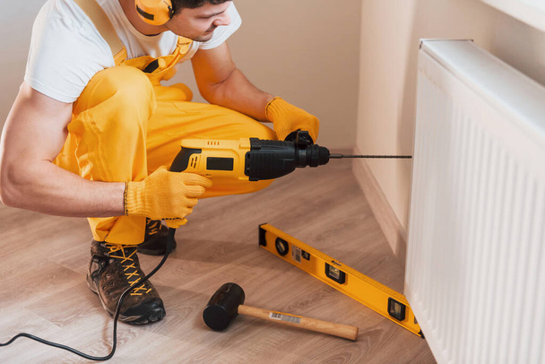 Handyman in yellow uniform works indoors by using hammer drill. House renovation conception. - 写真・画像