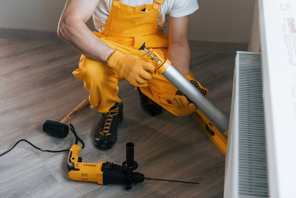 Handyman in yellow uniform works indoors with heat battery by using special tool. House renovation conception. - Fotoğraf, Görsel