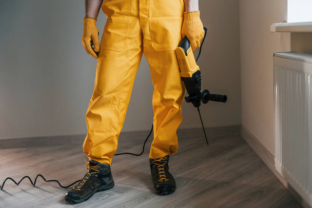 Handyman in yellow uniform with drill standing indoors. House renovation conception. - Фото, изображение