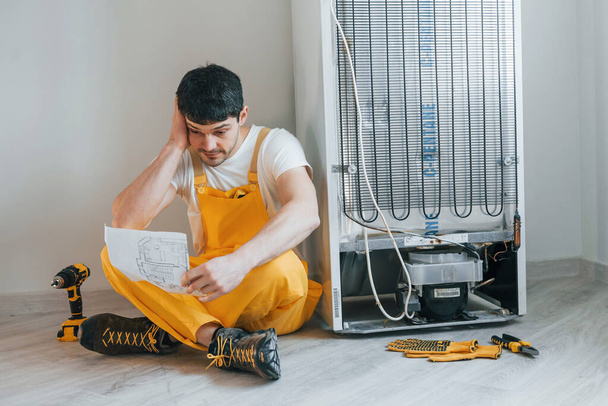 Confused handyman in yellow uniform reading manual for fridge indoors. House renovation conception. - Foto, Imagem