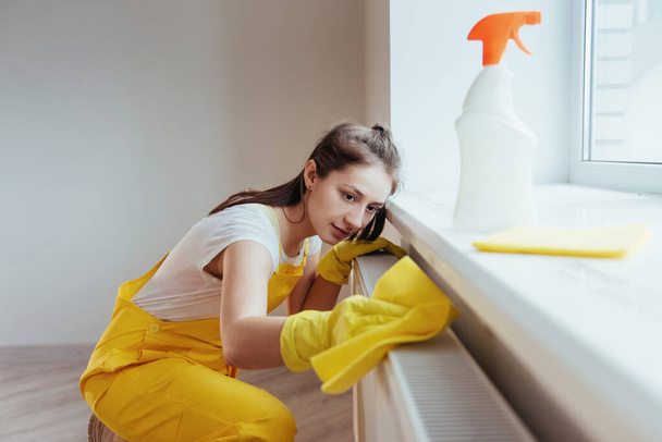 Ama de casa en uniforme amarillo trabaja con limpiador de ventanas y superficies en interiores. Concepto de renovación de casas. - Foto, imagen