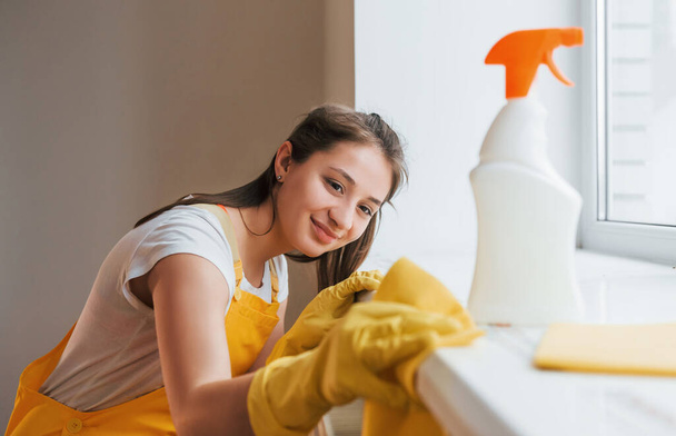 Housewife in yellow uniform works with window and surface cleaner indoors. House renovation conception. - Фото, изображение