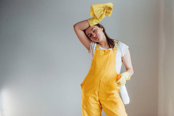 Femme au foyer fatiguée en uniforme jaune debout avec spray nettoyant pour fenêtres à l'intérieur. Conception de rénovation maison. - Photo, image