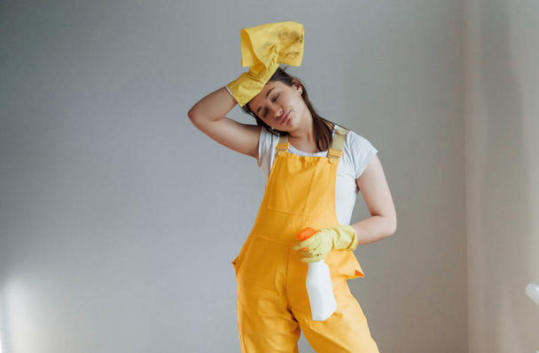 Уставшая домохозяйка в жёлтой форме стоит с моющим спреем для окон в помещении. Концепция реконструкции дома. - Фото, изображение