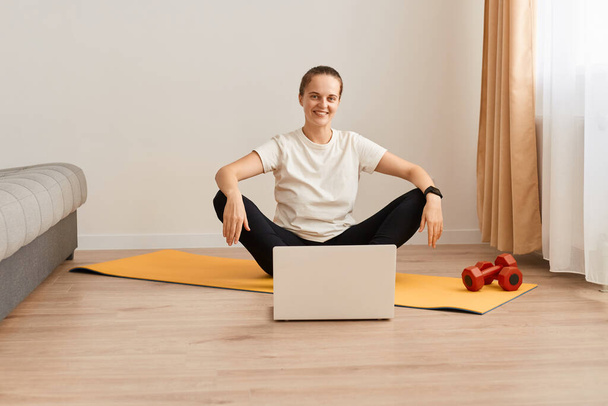 Indoor shot of female wearing white t shirt and black leggins sitting on mat with crossed arms, posing in front of laptop, watching tutorial video for yoga practice. - Valokuva, kuva
