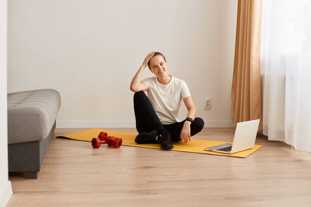 Portrait of tired young adult woman sitting on yoga mat with happy facial expressing after online training with trainer, female with sporty body wearing white t shirt and leggins. - Fotoğraf, Görsel