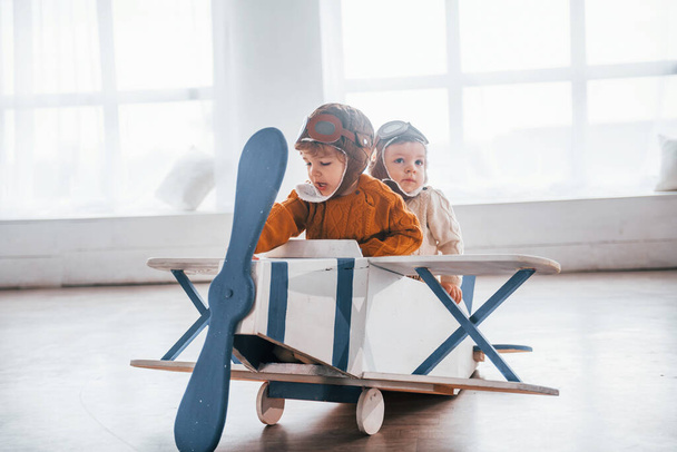 Two little boys in retro pilot uniform having fun with toy plane indoors. - Foto, afbeelding