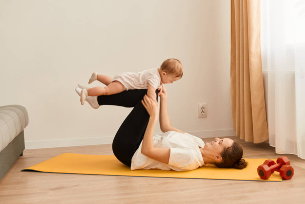 Attractive young mother doing sport exercises together with her infant baby, woman lying on floor on yoga mat and playing with her cute toddler kid while having workout at home. - Zdjęcie, obraz