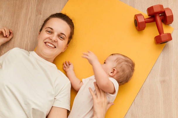 Top view of young mother with her baby daughter lying on the fitness mat during the exercise with dumbbells on the floor, resting, looking smiling at camera. - 写真・画像