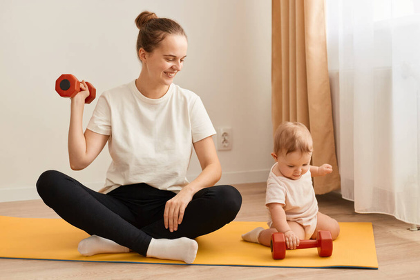 Indoor shot of smiling woman sitting on yoga mat with her baby daughter and training her arms, her biceps and triceps, doing sport exercises and looking after toddler kid. - Fotografie, Obrázek