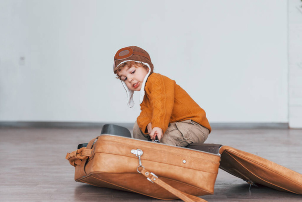 Little boys in retro pilot costume have fun and sitting in suitcase indoors at daytime. - Foto, imagen