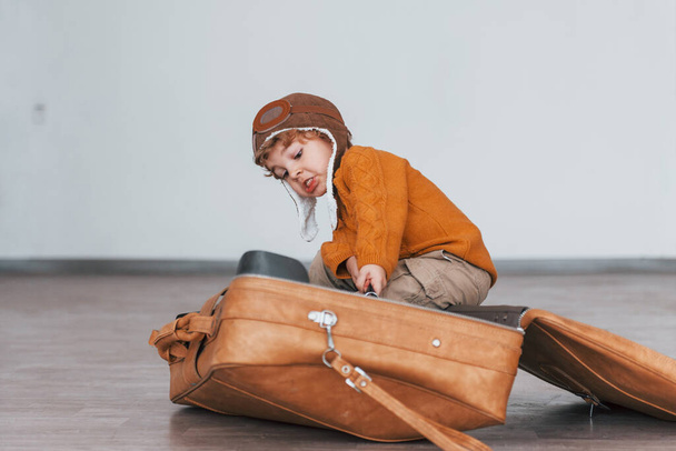 Little boys in retro pilot costume have fun and sitting in suitcase indoors at daytime. - Foto, Imagem