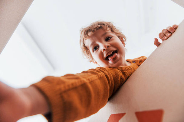 View from below of little boy with curly hair that have fun with paper box indoors. - Foto, Bild