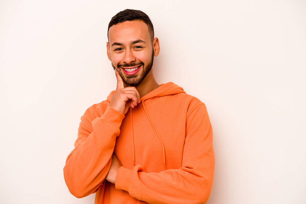 Young hispanic man isolated on white background smiling happy and confident, touching chin with hand. - 写真・画像