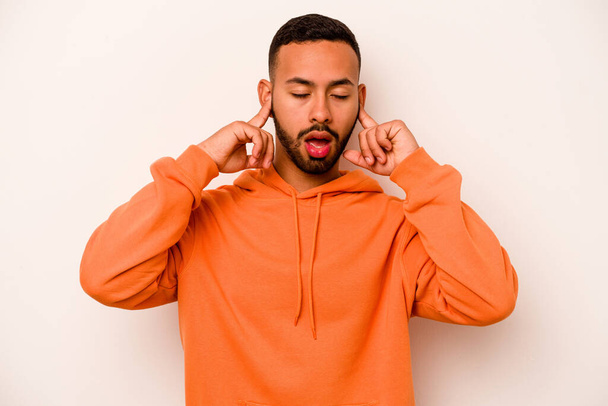 Young hispanic man isolated on white background covering ears with fingers, stressed and desperate by a loudly ambient. - Фото, изображение