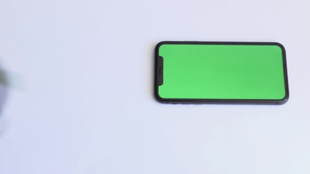 Τοποθετήστε για τσάι πόσιμο με smartphone σε λευκό τραπέζι με πράσινη οθόνη. Πράσινο τσάι σε ποτήρι. Κοντινό πλάνο του κινητού τηλεφώνου σε λευκό τραπέζι με chroma κλειδί, πράσινο τηλέφωνο οθόνη και πάνω όψη - Πλάνα, βίντεο