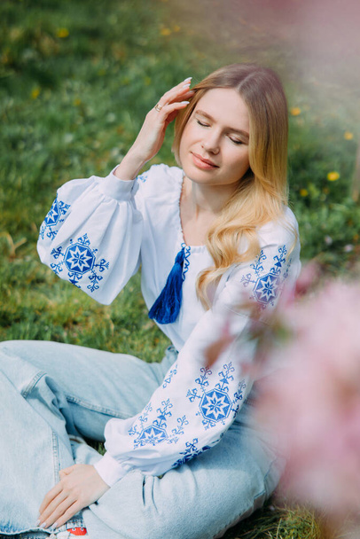Νεαρή ξανθιά γυναίκα στηρίζεται στο γκαζόν στον κήπο ντυμένος στην ουκρανική κεντημένο πουκάμισο κοντά στο ανθισμένο δέντρο sakura. Κλείσιμο. - Φωτογραφία, εικόνα