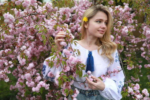Portrait de jeune femme blonde en fleurs jardin sakura habillé en chemise brodée ukrainienne avec fleur de pissenlit. - Photo, image