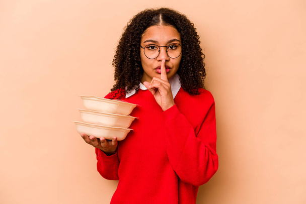 Young African American woman holding tupperware isolated on beige background keeping a secret or asking for silence. - Foto, afbeelding