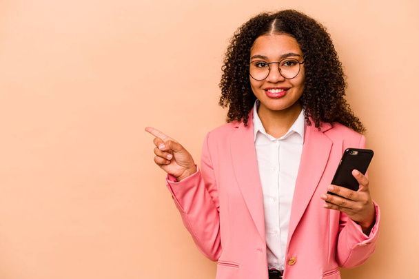 Young African American woman holding mobile phone isolated on beige background smiling and pointing aside, showing something at blank space. - Foto, imagen