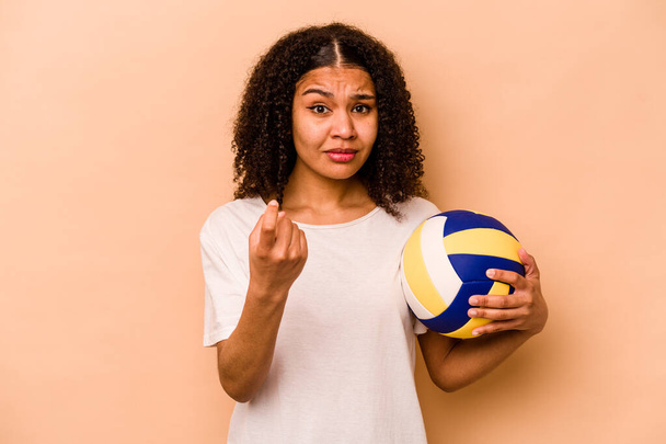 Молодая афроамериканка, играющая в волейбол на бежевом фоне, тычет пальцем в тебя пальцем, словно приглашая подойти ближе.. - Фото, изображение