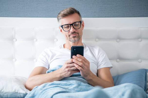cheerful mature man in bed chatting on phone wearing eyeglasses - 写真・画像