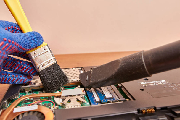 A személy egy kefével és egy porszívóval tisztítja a laptop hűtőrendszerét. PC gondozási és javítási koncepció. - Fotó, kép