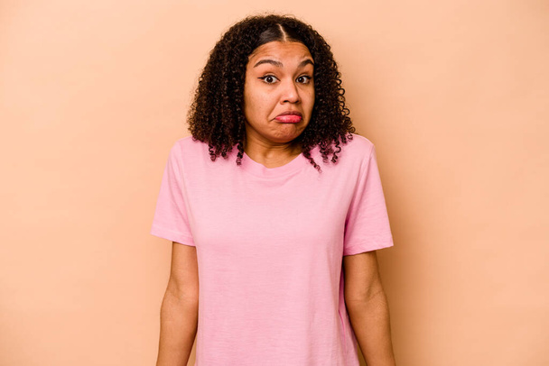 Young African American woman isolated on beige background shrugs shoulders and open eyes confused. - Photo, Image
